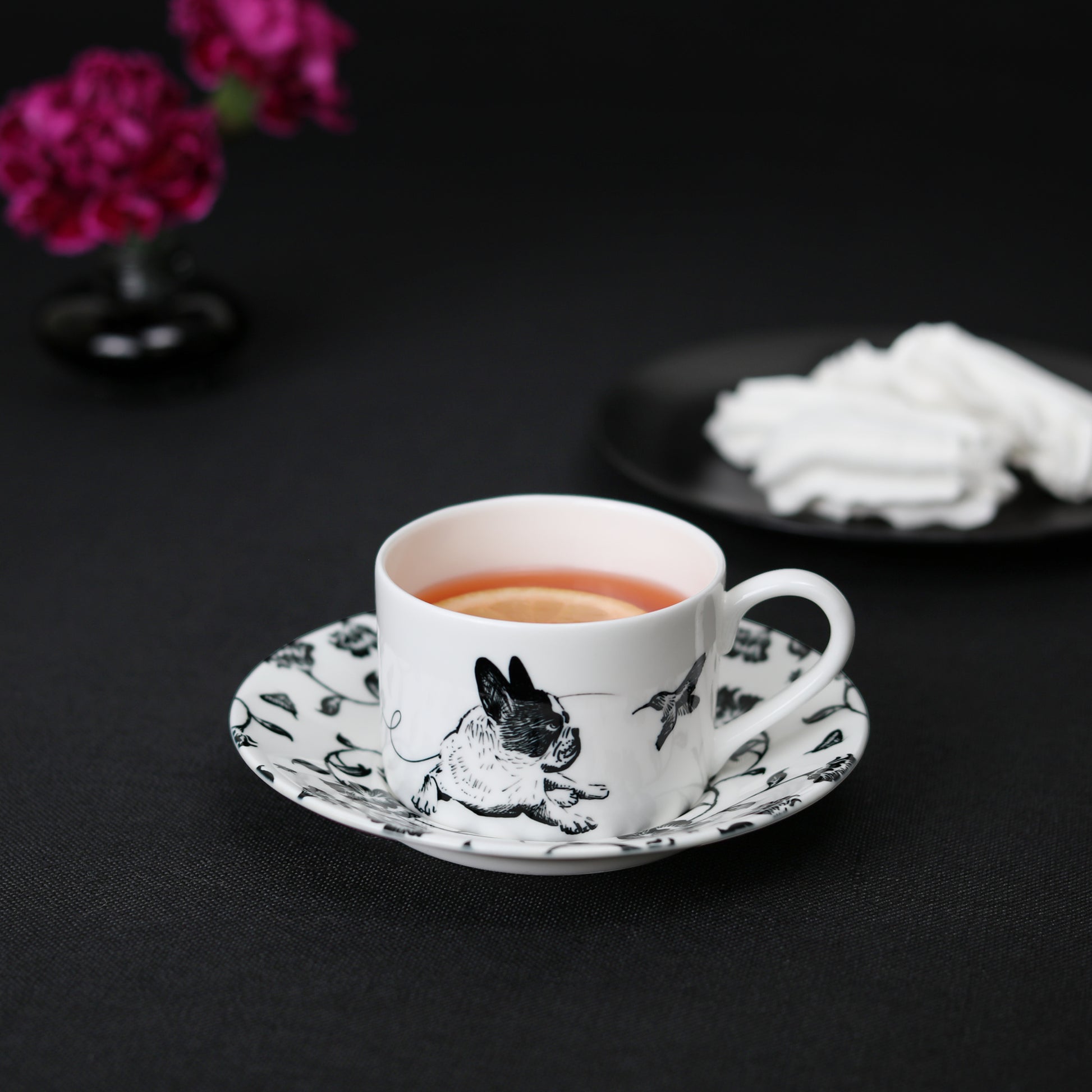 A black and white cup and saucer featuring a french bulldog design, elegantly displayed on a table with tea, meringue cookies, and flowers.