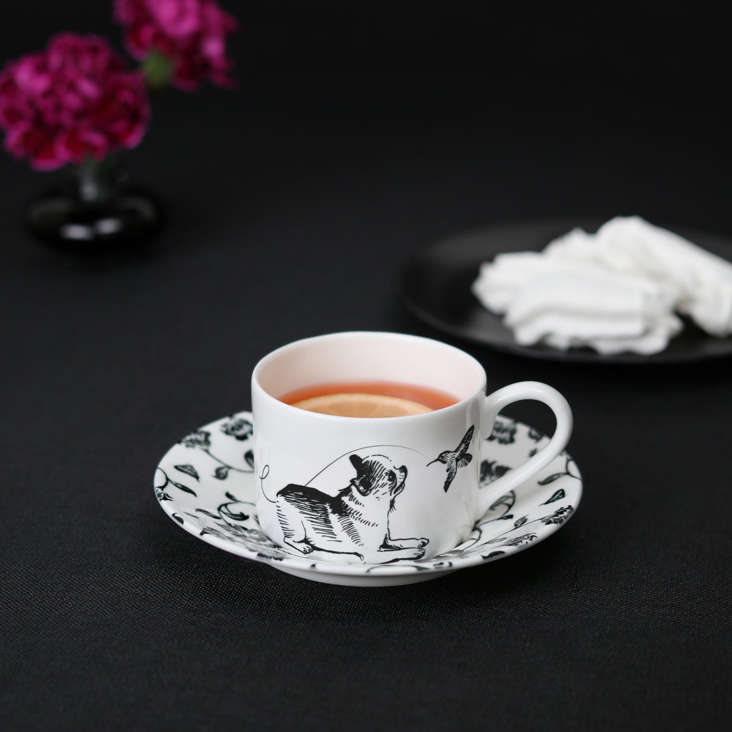 A black and white cup and saucer featuring a chihuahua design, elegantly displayed on a table with tea, meringue cookies, and flowers.