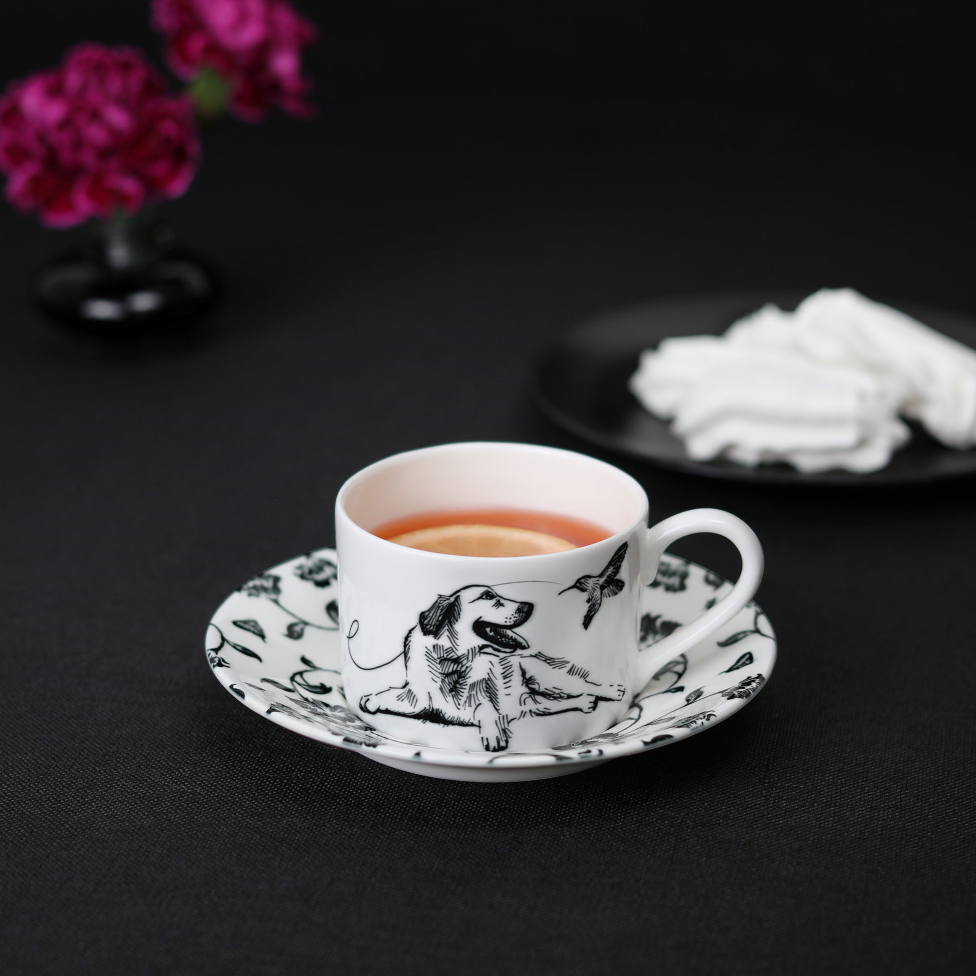 A black and white cup and saucer featuring a labrador retriever design, elegantly displayed on a table with tea, meringue cookies, and flowers.