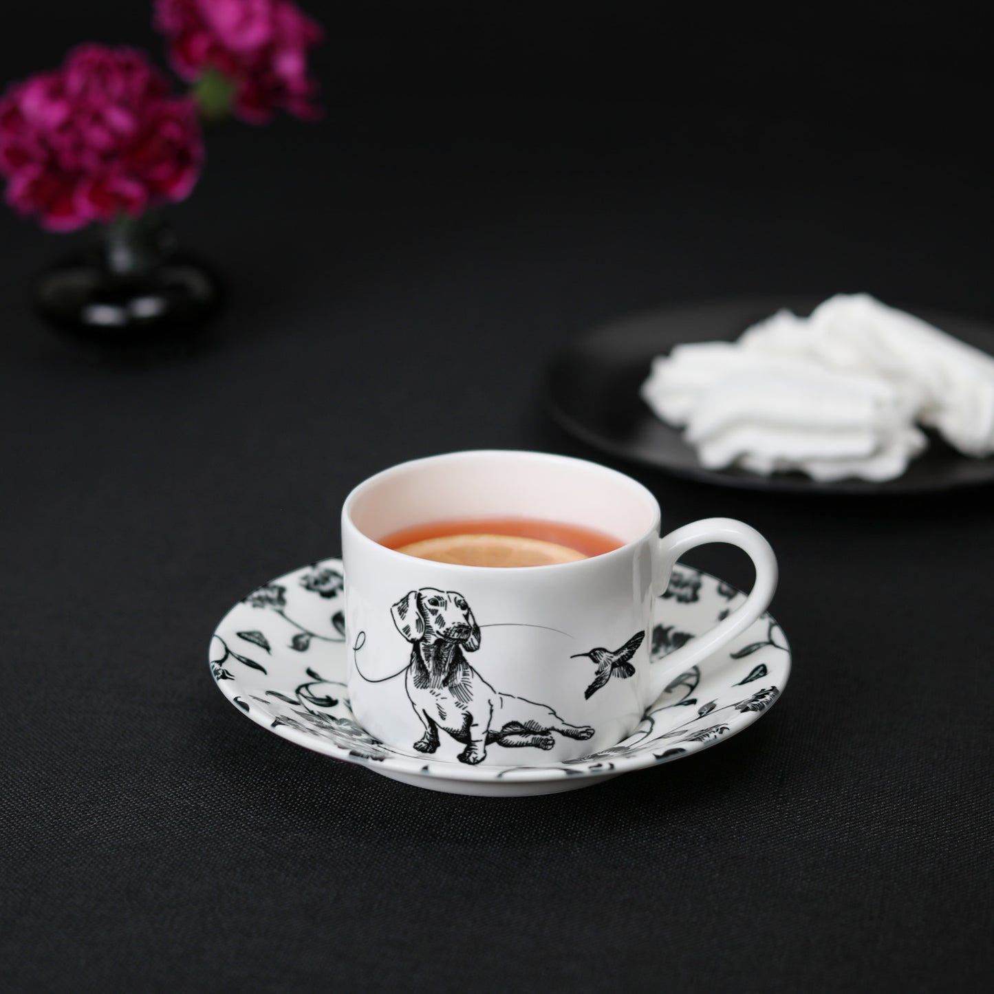 A black and white cup and saucer featuring a dachshund design, elegantly displayed on a table with tea, meringue cookies, and flowers.