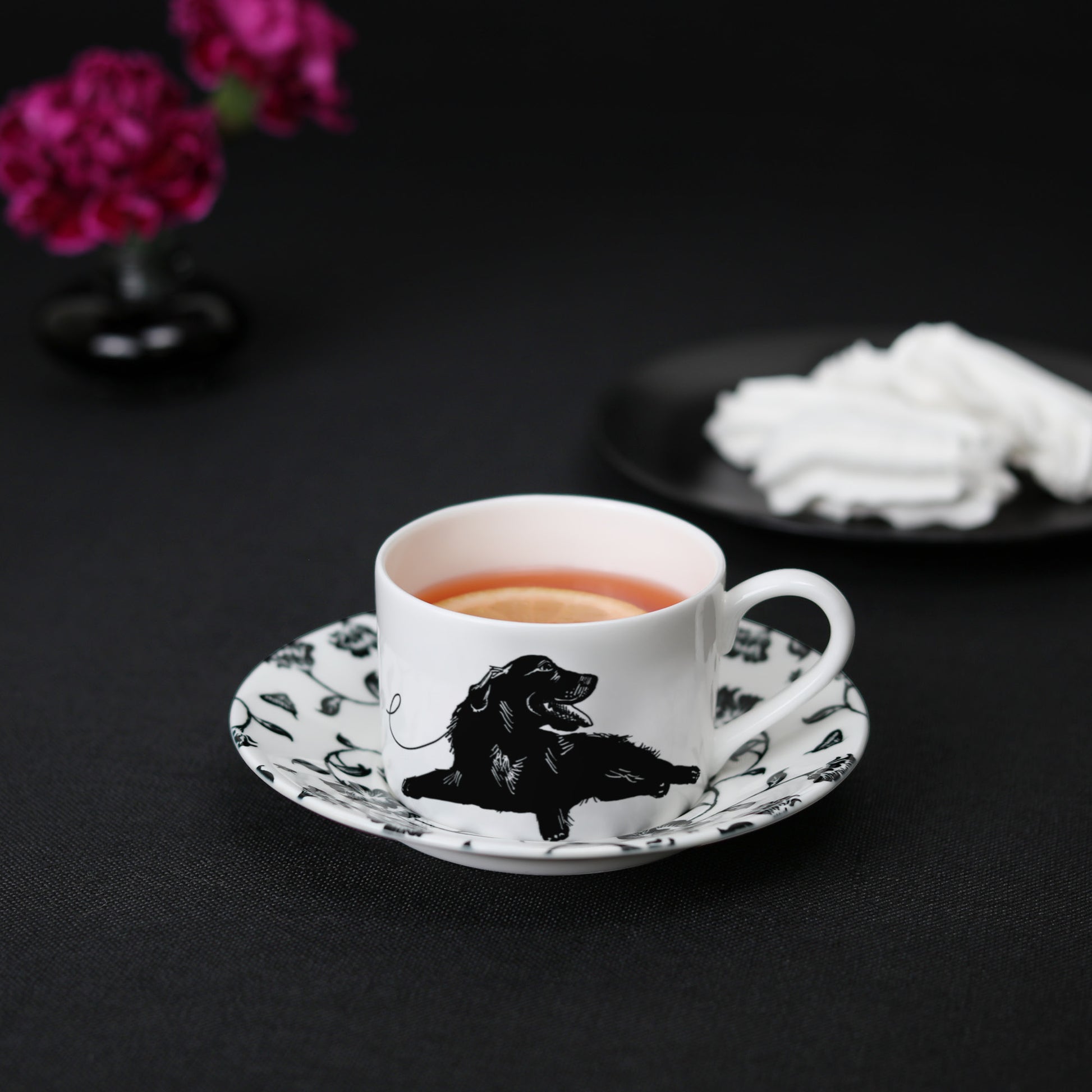 A black and white cup and saucer featuring a black labrador design, elegantly displayed on a table with tea, meringue cookies, and flowers.