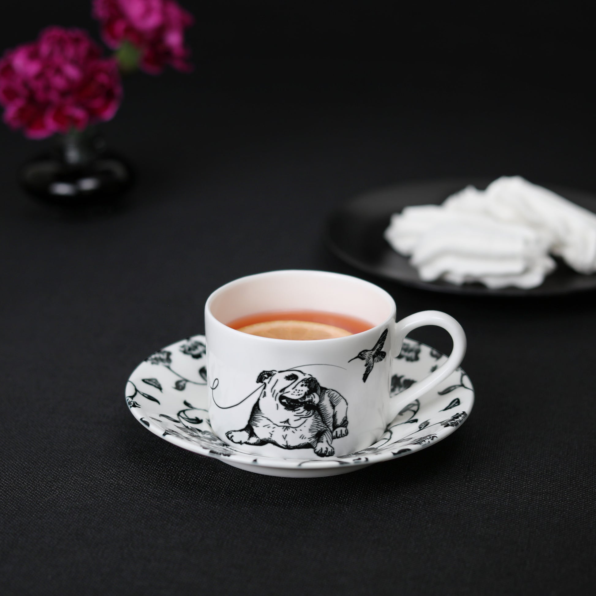 A black and white cup and saucer featuring a bulldog design, elegantly displayed on a table with tea, meringue cookies, and flowers.